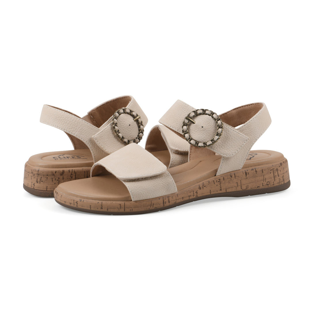Angle view of a pair of cream sandals with double adjustable straps, circular buckle embellishment, and low cork print wedge 