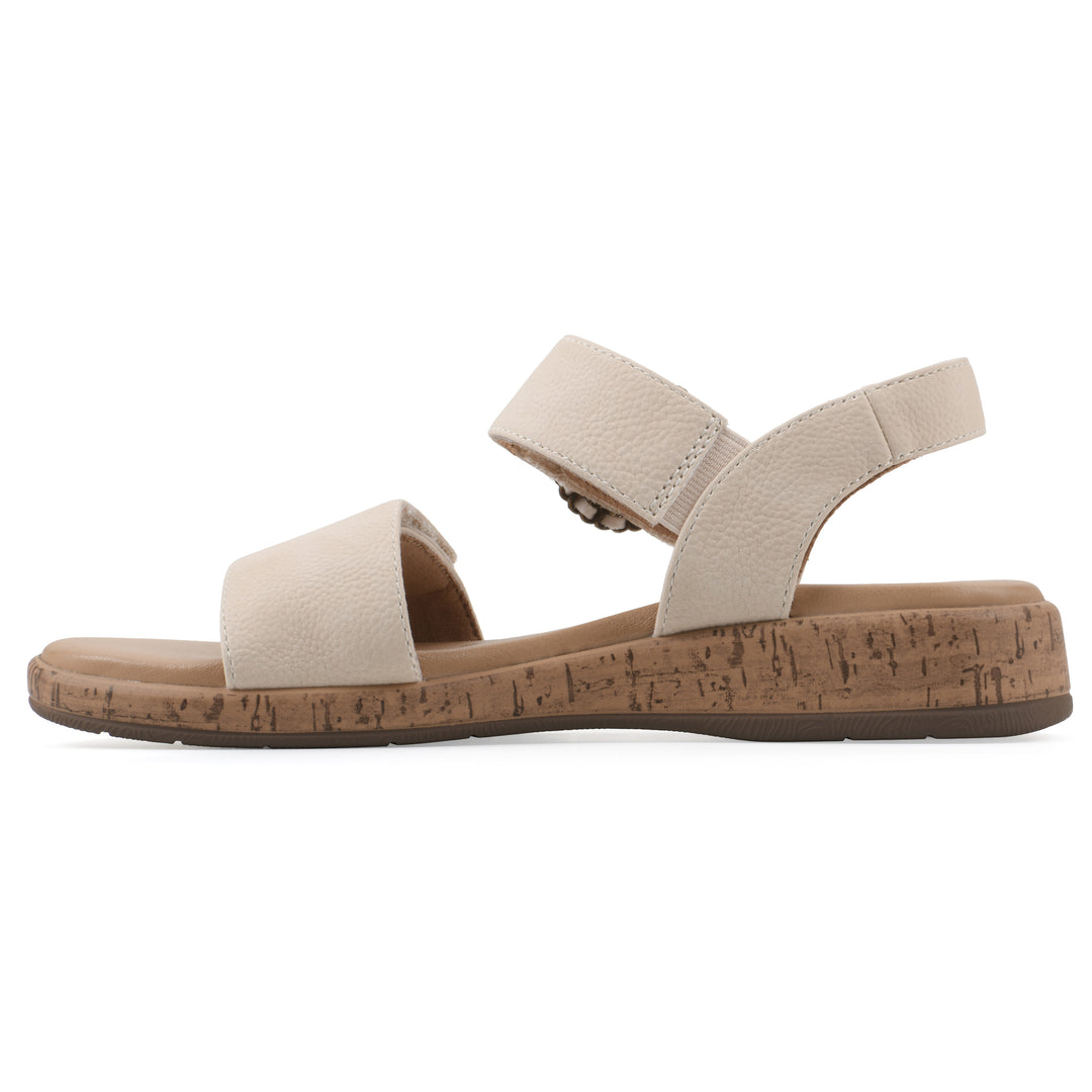 Side view of a cream sandal with double adjustable straps, circular buckle embellishment, and low cork print wedge 