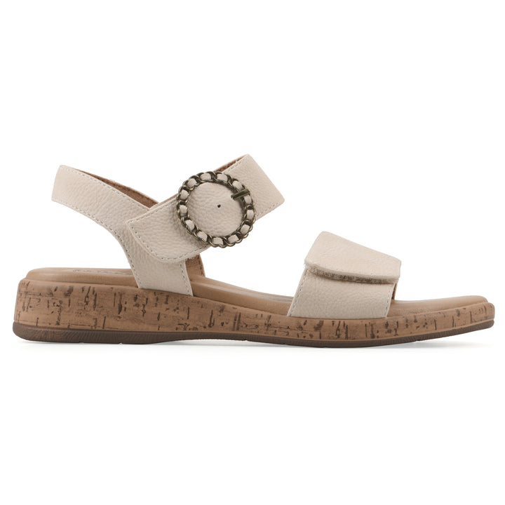 Side view of a cream sandal with double adjustable straps, circular buckle embellishment, and low cork print wedge 