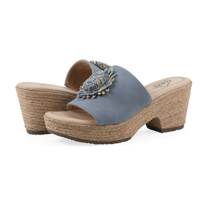 Angle view of a pair of blue, backless wedge sandals with centered sunflower embellishment, and braided rope style heel