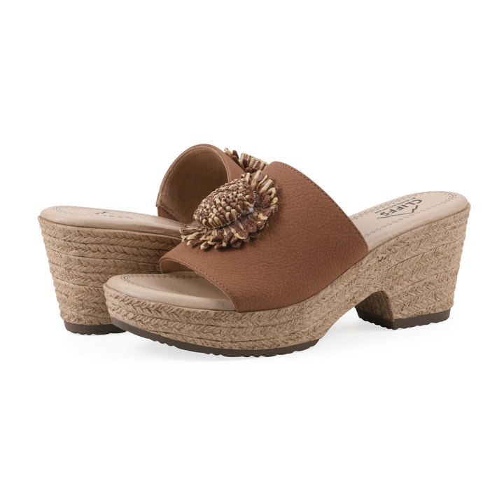 Angle view of a pair of brown, backless wedge sandals with centered sunflower embellishment, and braided rope style heel