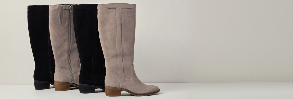 Four tall knee-high boots in black and taupe suede, arranged in a row against a minimalist light gray background.