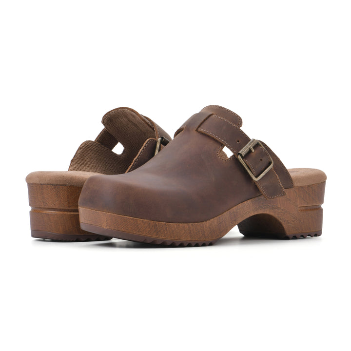 Angle view of a pair of backless brown leather clogs with adjustable buckle and wood style heel